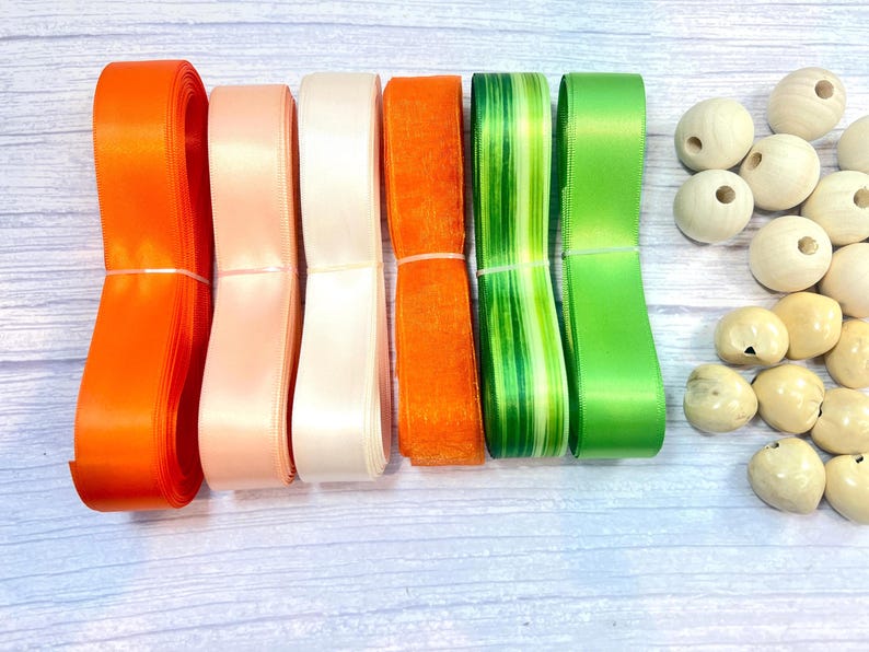 Peut inclure: Un assortiment de rubans color&eacute;s et de perles en bois. Les rubans sont dans les tons orange, rose, blanc, vert et un motif ray&eacute; vert et blanc. Les perles en bois sont marron clair et ont des trous.
