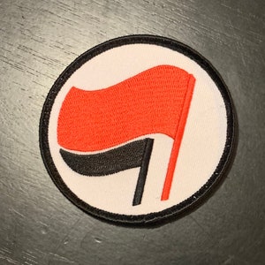 May include: A white circle patch with a black and red flag design. The flag is red with a black stripe and a white pole.