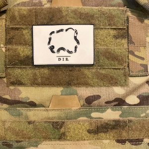 May include: A white patch with a black outline of a stylized figure and the text "D.I.B."  The patch is attached to a camouflage fabric background.