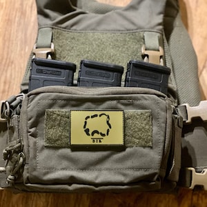 May include: A green tactical vest with three black magazine pouches and a small green pouch with a gold patch that says "DIR" with a stylized image of a wolf's head.