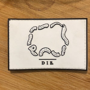 Könnte beinhalten: Ein rechteckiger weißer Aufnäher mit schwarzem Rand. Der Aufnäher zeigt eine schwarz-weiße Grafik eines Kreises aus segmentierten Formen, mit dem Wort "DIE" darunter in Schwarz gedruckt.