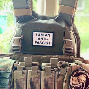 May include: A tactical plate carrier vest in a camouflage pattern featuring a rectangular white patch with black text that reads I AM AN ANTI-FASCIST. The vest includes multiple utility pouches, adjustable shoulder straps, and a circular patch.