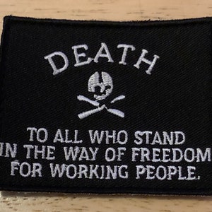 Könnte beinhalten: Ein rechteckiger, schwarzer, bestickter Aufnäher mit weißem Text, der DEATH TO ALL WHO STAND IN THE WAY OF FREEDOM FOR WORKING PEOPLE lautet. Über dem Text befindet sich ein weiß gesticktes Totenkopf-Symbol mit gekreuzten Knochen.