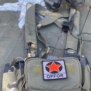 May include: A green tactical vest with a red star patch that says "OPFOR" and a first aid pouch with a red cross. The vest has a magazine pouch and a radio attached to it.