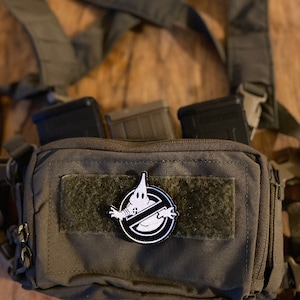 Könnte beinhalten: Eine grüne taktische Tasche mit einem schwarz-weißen Patch, auf dem ein Geist mit einem Verbotszeichen zu sehen ist. Die Tasche hat drei schwarze Magazin-Taschen auf der Vorderseite.