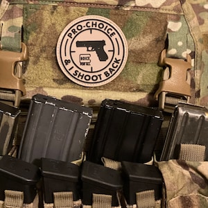 Könnte beinhalten: Eine taktische Tarnweste mit einem runden beigen Aufnäher, der eine schwarze Pistole und den Text PRO-CHOICE & I SHOOT BACK sowie HOICE FOR ALL zeigt. Die Weste enthält mehrere schwarze Gewehr- und Pistolenmagazine in integrierten Taschen.