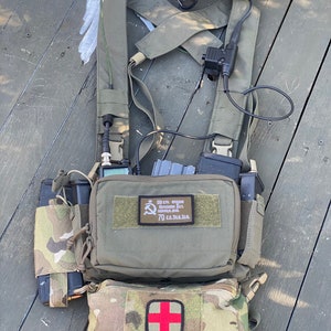 May include: A green tactical chest rig with a variety of pouches and accessories. The rig has a large main pouch with a patch that reads "15 cта. 90мм.  Дымовая граната. 70 с.м. 3.1.1.6. ". The rig also has a smaller pouch with a red cross on it.