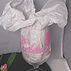 Puede incluir: Una copa de vino de vidrio transparente con una calcomanía de vinilo rosa que dice "Relax" y una silueta de copa de vino rosa. La copa está envuelta en papel de seda blanco.