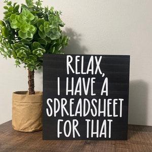Puede incluir: Un letrero de madera negro con texto blanco que dice "Relax, I have a spreadsheet for that". El letrero est&aacute; sobre una superficie de madera delante de una planta verde en maceta.