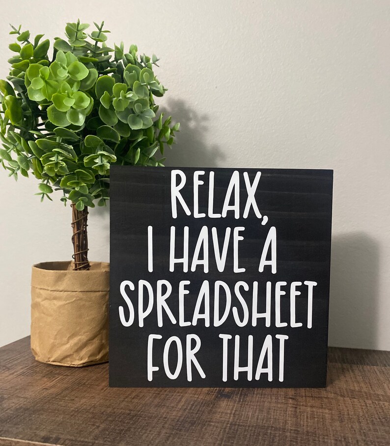 Puede incluir: Un letrero de madera negro con texto blanco que dice "Relax, I have a spreadsheet for that". El letrero est&aacute; sobre una superficie de madera delante de una planta verde en maceta.