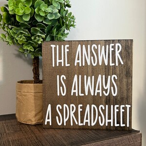 May include: A wooden sign with the text "THE ANSWER IS ALWAYS A SPREADSHEET" in white lettering on a dark brown background.