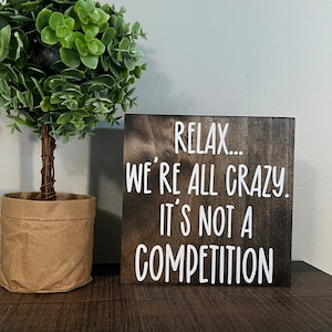 May include: A wooden sign with the text "Relax... We're all crazy. It's not a competition." in white lettering on a dark brown background.