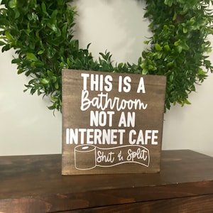 May include: A brown wooden sign with white text that reads "This is a bathroom not an internet cafe" with a toilet paper roll graphic and the text "Shit & Split" underneath.