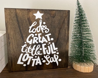 May include: A wooden sign with the words "looks GREAT little FULL LOTTA sap" in a Christmas tree shape. The sign is dark brown with white lettering and a white star at the top. A small green Christmas tree is to the right.