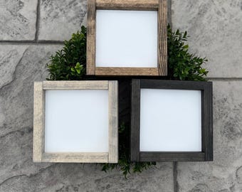 May include: Three square wooden frames in varying shades of brown, gray, and black, each with a white interior. The frames are arranged in a triangular pattern and are surrounded by green foliage, creating a rustic aesthetic.