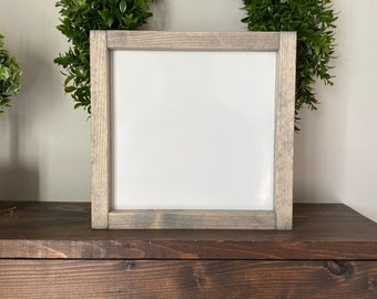 May include: A white sign with a gray wood frame sits on a dark brown wooden shelf. A green wreath is behind the sign.