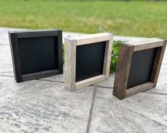 May include: Three small, square wooden chalkboards with black surfaces. The frames are in varying shades of gray and brown, with visible wood grain. The chalkboards are leaning against a textured gray surface, with green grass in the background.