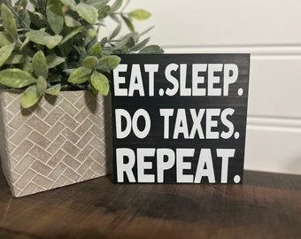 Könnte beinhalten: Schwarzes Holzschild mit weißem Text, der "EAT. SLEEP. DO TAXES. REPEAT." lautet.