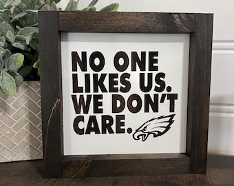 Könnte beinhalten: Quadratisches Holzschild mit einem dunkelbraunen Rahmen. Der weiße Innenraum zeigt den Text "NO ONE LIKES US. WE DON'T CARE." in fetten schwarzen Buchstaben sowie ein schwarzes Adlerlogo. Das Schild wird auf einer Holzoberfläche angezeigt.