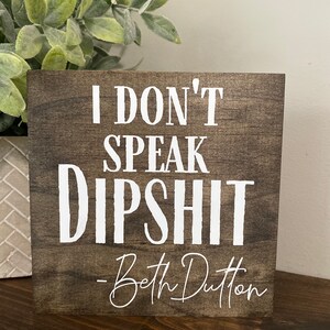 May include: A wooden sign with the text "I don't speak dipshit - Beth Dutton" in white lettering.
