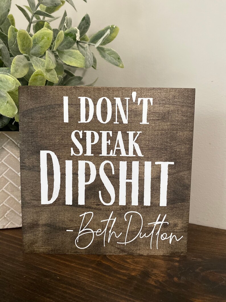May include: A wooden sign with the text "I don't speak dipshit - Beth Dutton" in white lettering.