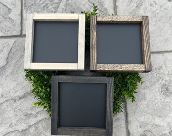 May include: Three small square chalkboards with wooden frames in varying shades of gray and brown. The chalkboards are arranged on a bed of green foliage, creating a rustic and decorative display. The frames have a distressed finish.