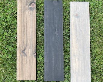 May include: Three wooden boards, each stained a different color: brown, black, and gray. The boards are arranged side-by-side on a bed of green grass.