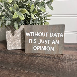 May include: A wooden sign with the text "WITHOUT DATA IT'S JUST AN OPINION" in white lettering on a gray background. The sign is sitting on a wooden surface.