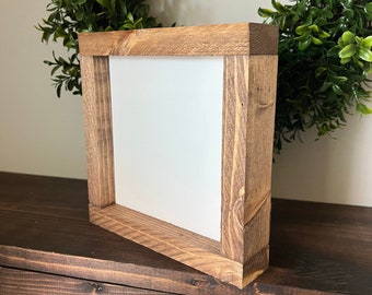 May include: A small, square wooden frame with a white background. The frame is made of natural wood with a dark brown stain. The frame is sitting on a wooden surface.