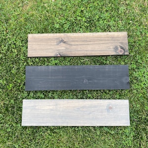 May include: Three wooden planks, one stained a dark brown, one stained black, and one stained a light gray. The planks are arranged vertically on a green grassy surface.