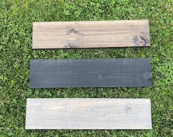 May include: Three rectangular wooden planks in varying shades of stain: light brown, black, and light gray. Each plank has a visible wood grain and a knot. The planks are arranged horizontally on a bed of green grass.