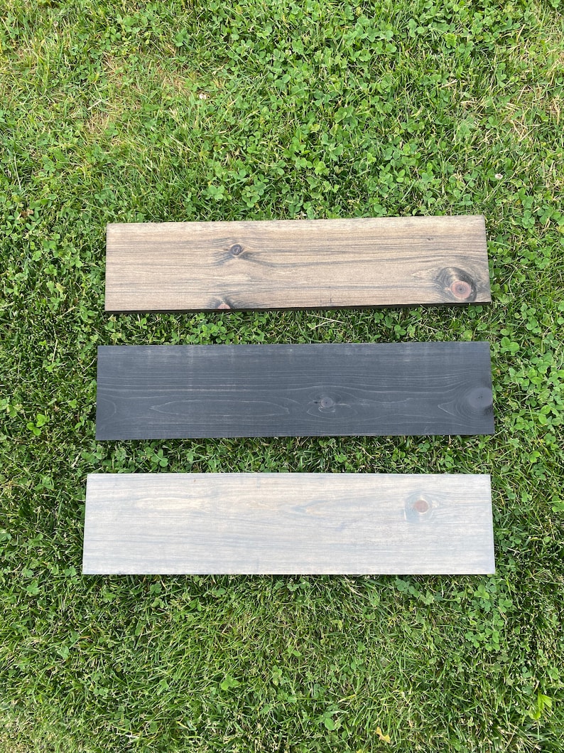 May include: Three wooden planks, one stained a dark brown, one stained black, and one stained a light gray. The planks are arranged vertically on a green grassy surface.