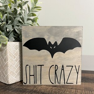 May include: A gray wooden sign with a black bat silhouette and the text "SHIT CRAZY" in black lettering.