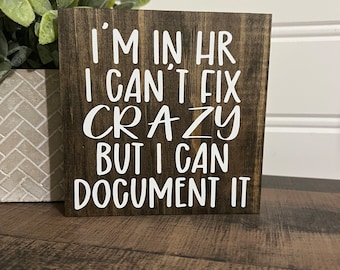 May include: A wooden sign with the text "I'm in HR I can't fix crazy but I can document it" in white lettering.