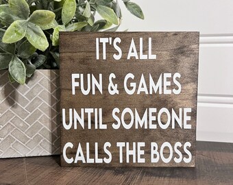 May include: A wooden sign with the text "It's all fun & games until someone calls the boss" in white lettering.