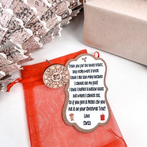 May include: A red organza bag with a wooden tag attached. The tag has a brown background with a white cloud shape. The text on the tag reads: "Thank you for the lovely snacks, they really were a treat. I think I ate too many because I cannot see my feet! I think I popped a button loose but where I cannot see, so if you find it please can you put it on your Christmas Tree? Love Santa".