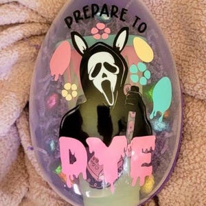 Può includere: Un contenitore viola a forma di uovo con la scritta "PREPARE TO DYE" in rosa. Il design presenta una figura nera con una maschera di Scream con orecchie da coniglio, che tiene un coltello e gocce di vernice colorate.