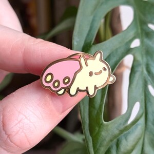 May include: A gold-colored enamel pin featuring a cartoon-style creature with a pink shell and yellow body. The pin is held in front of a green plant with large, patterned leaves. The pin has a gold outline.