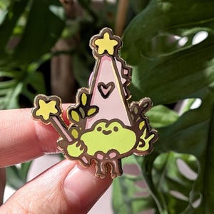 May include: A whimsical enamel pin featuring a cartoon frog wearing a party hat with a star and heart design. The pin has a gold outline and a yellow star wand. The frog is light green, and the hat is pink.