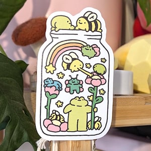 Op de afbeelding: Een grillige sticker met een pot vol cartoonbijen, kikkers, bloemen en een regenboog. De sticker heeft een witte rand en is gevuld met pastelkleuren. De pot is gevuld met een verscheidenheid aan schattige wezens en planten.