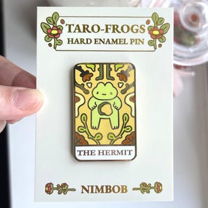 May include: A hard enamel pin featuring a frog tarot card design. The pin has a yellow background with a green frog illustration and the words "THE HERMIT". The pin is on a white card with the text "TARO-FROGS" and "NIMBOB".