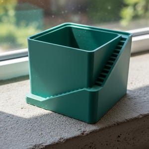 Könnte beinhalten: Ein türkisfarbener, quadratischer Blumentopf mit einem Treppendesign an einer Seite. Der Topf steht auf einem passenden Sockel auf einer Fensterbank aus Beton. Im Hintergrund ist ein Fenster mit verschwommenem Blick auf Grün zu sehen. Ca. 10 cm x 10 cm.