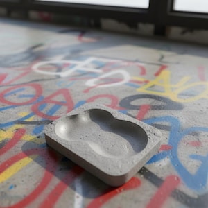Könnte beinhalten: Ein kleines, rechteckiges, graues Beton-Tablett mit zwei abgerundeten Vertiefungen. Das Tablett steht auf einer Betonoberfläche, die mit farbenfrohen Graffiti bedeckt ist, darunter rote, blaue, gelbe und weiße Sprühfarbe. Der Hintergrund umfasst ein Fenster.