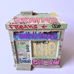 May include: A concrete miniature building with graffiti art. The roof features the word "BRAINS" in pink, and the sides are covered in colorful tags and phrases like "FREAKS" and "CHEW". The building has a small, rectangular chimney.