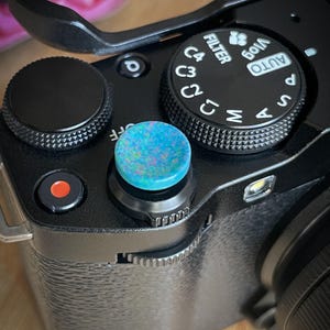 Pode incluir: Close-up de uma câmera digital preta com um botão colorido e iridescente. A câmera possui vários mostradores e botões, incluindo um seletor de modo com configurações como "AUTO" e "FILTER". O botão é uma mistura vibrante de azuis, verdes e rosas.