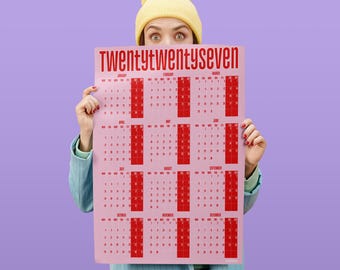 May include: A wall calendar for the year twenty twenty-seven featuring a light pink background with red text and grid lines. The calendar displays all twelve months of the year, with the title twenty twenty-seven printed in large red letters at the top.