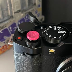 May include: Close-up of a black camera body with a pink textured button. The camera has several dials and buttons, including a mode dial with settings like AUTO and M. The camera also has a small red button.