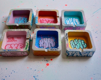 May include: Six square concrete ashtrays featuring colorful graffiti-style tags and lettering. Each ashtray has a unique interior color including pink, orange, light blue, and yellow, with hand-painted text like HYPE and LOVE YOUR FREEDOM.