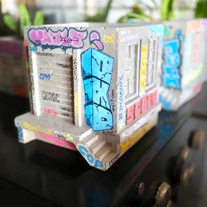 Könnte beinhalten: Beton-Pflanzkasten mit Graffiti-Kunst. Der graue, rechteckige Pflanzkasten weist blau, rosa und gelb gesprühte Designs auf. Der Pflanzkasten hat ein Gebäude-Fassaden-Design mit einem blauen Graffiti-Tag an der Seite.