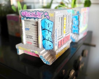 May include: Concrete planter box with graffiti art. The grey rectangular planter features blue, pink, and yellow spray-painted designs. The planter has a building facade design with a blue graffiti tag on the side.